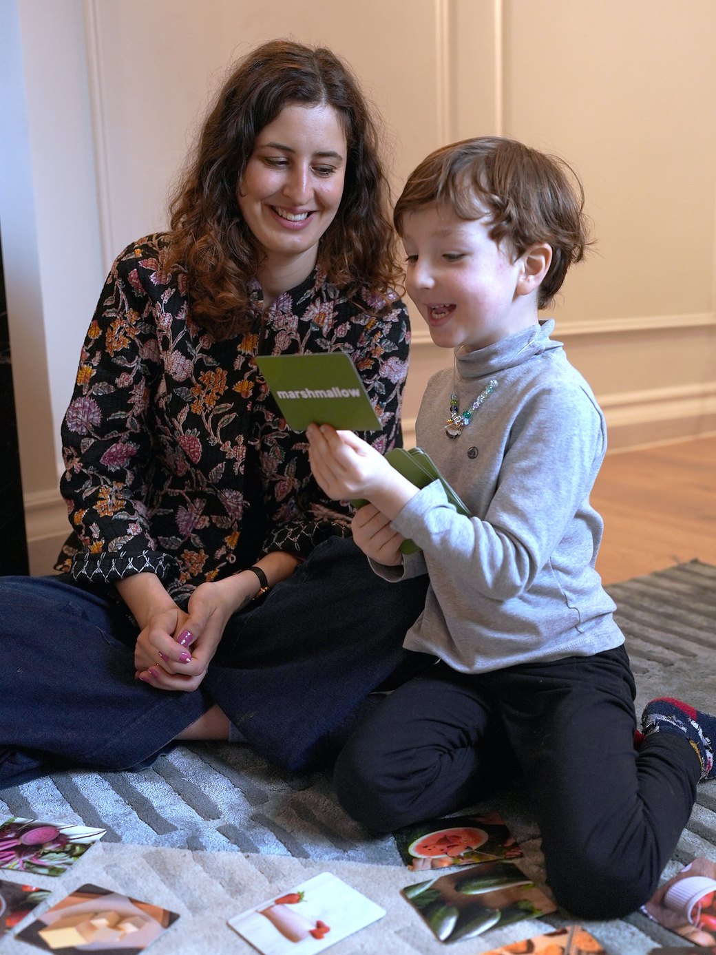 Beatrice smiling as a boy reads a 'marshmallow' word card, with picture cards on the rug